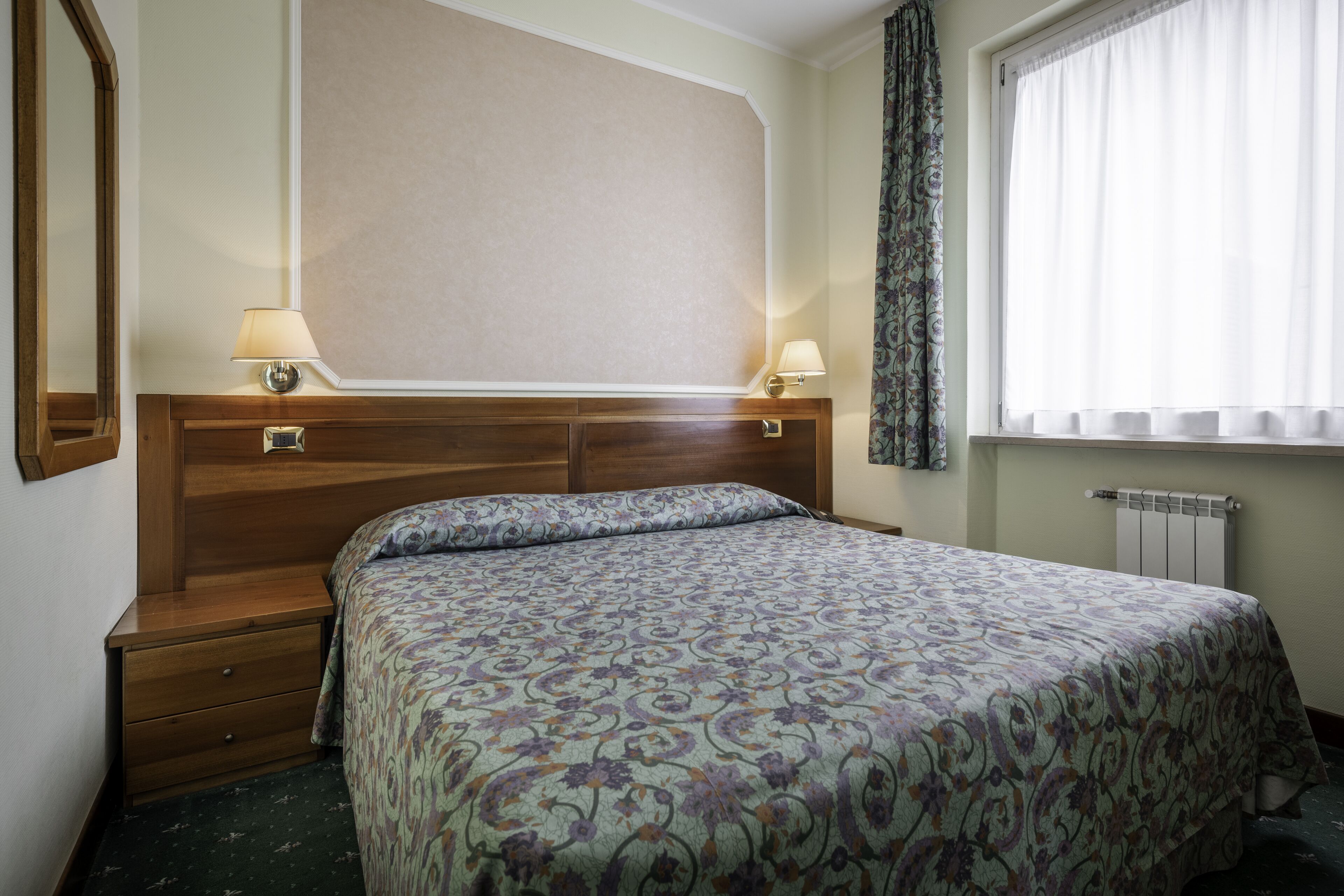 A hotel room with a wooden headboard, two bedside tables with lamps, a patterned bedspread, a window with sheer curtains, and a radiator below.
