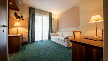Una camera d'albergo con tappeto verde, un divano, una scrivania, due lampade, una finestra con tende e un fioriero a parete.