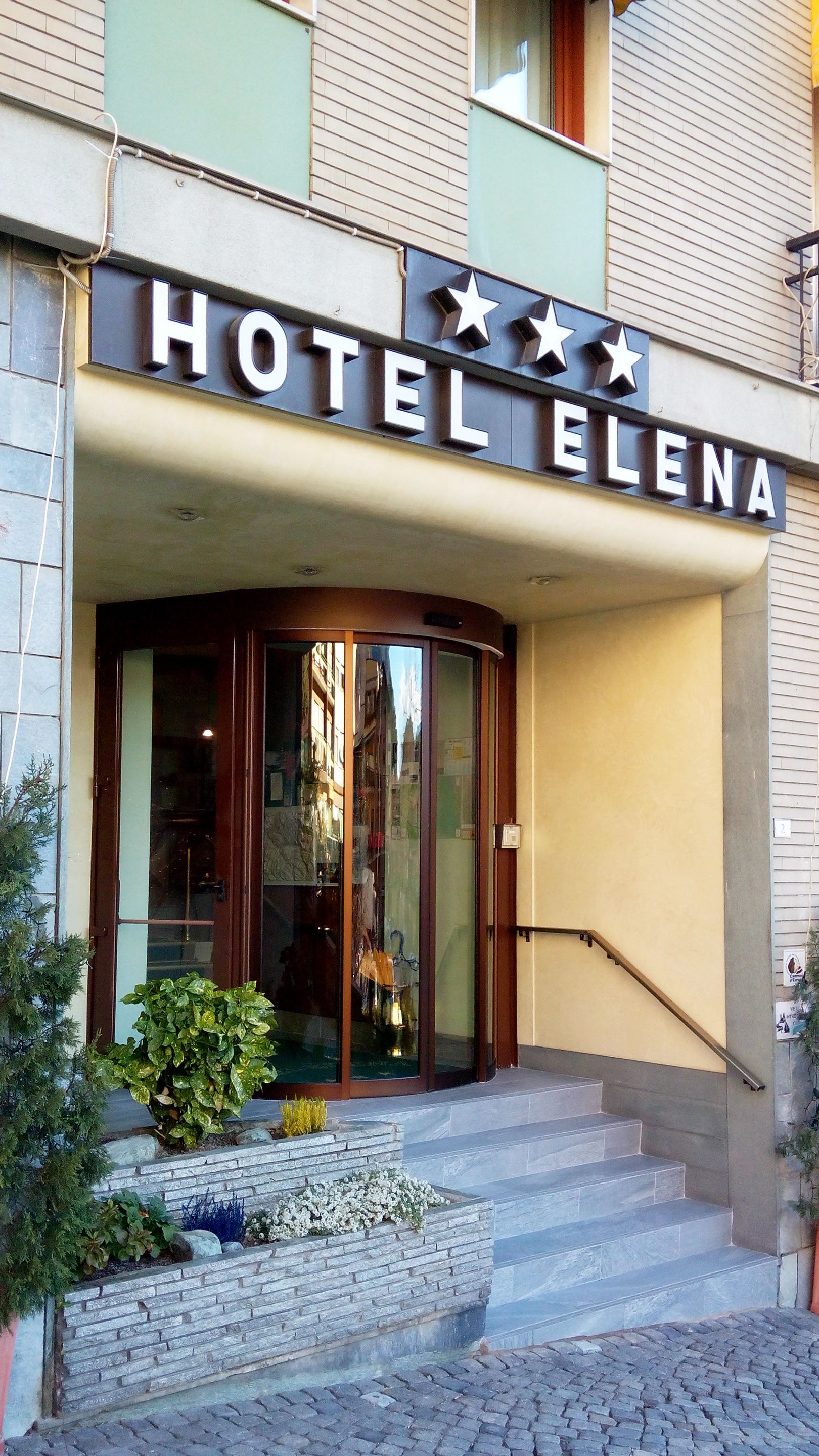 L’entrée de l’hôtel Elena se compose d’un panneau indicateur, de portes vitrées, d’un escalier et d’un petit jardin.