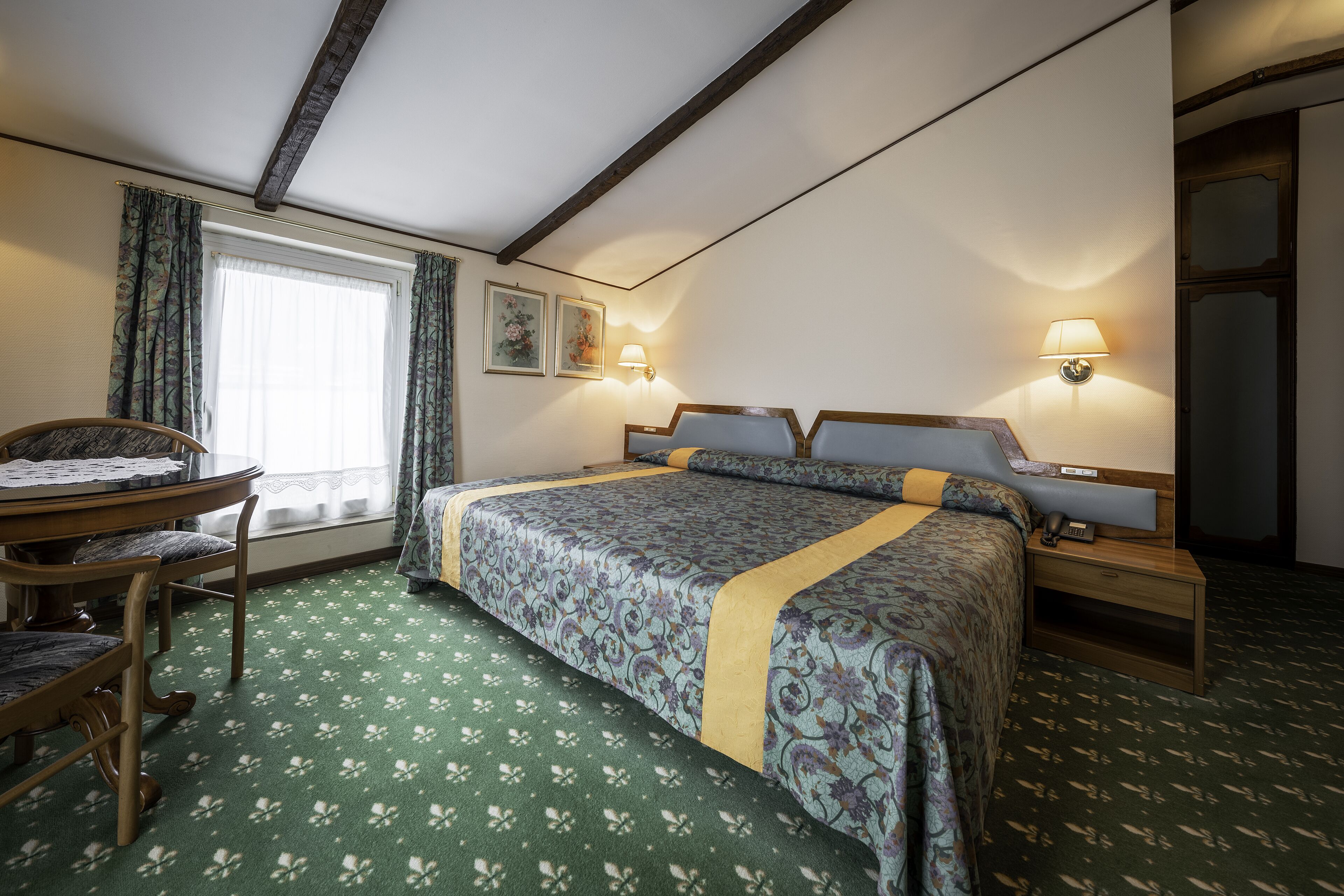 A hotel room with a bed, a desk, a chair, a window with curtains, and two wall-mounted lamps.