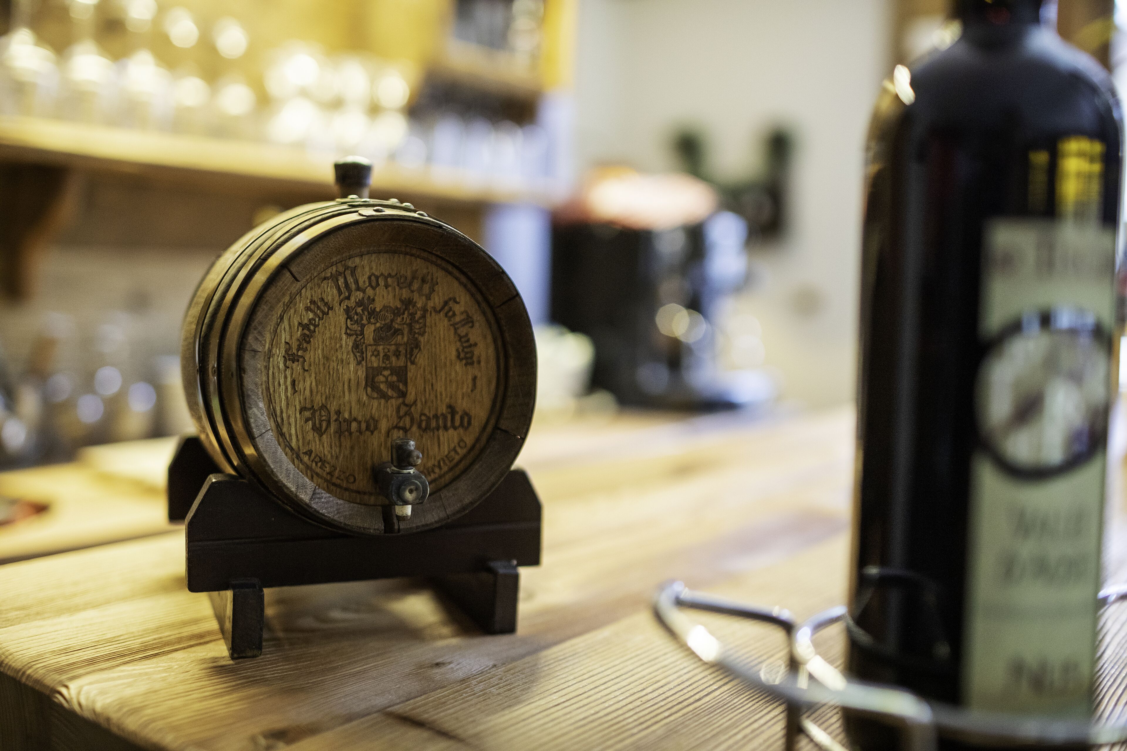 A wooden barrel with a tap on a wooden table, a bottle of wine, and a pair of glasses.