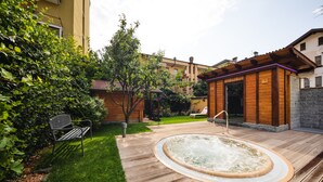 Une terrasse en bois avec un jacuzzi, un banc et un petit espace jardin agrémenté d’un arbre et d’arbustes.