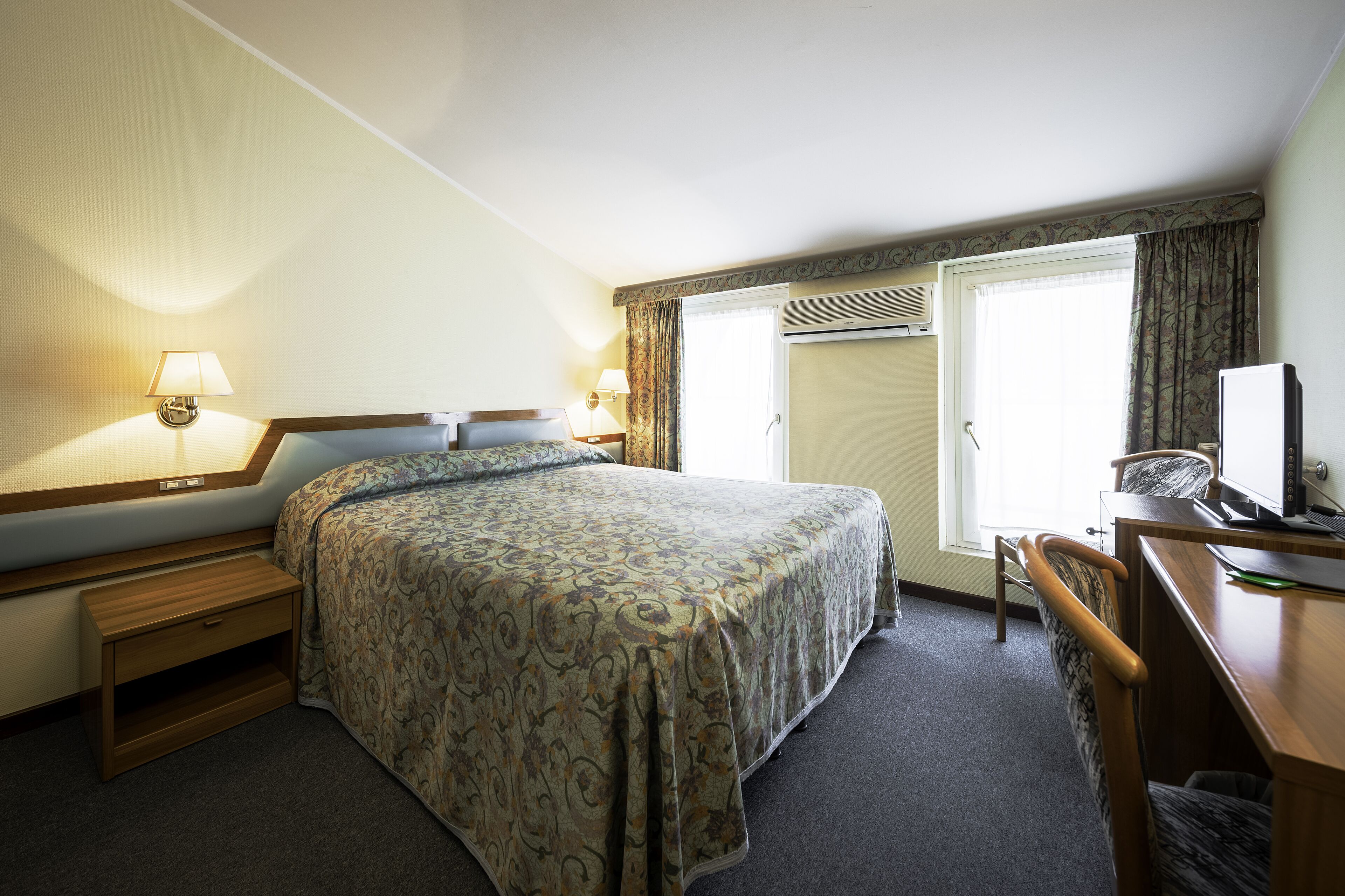 Habitación de hotel con cama, mesitas de noche, escritorio con ordenador y ventana con cortinas.