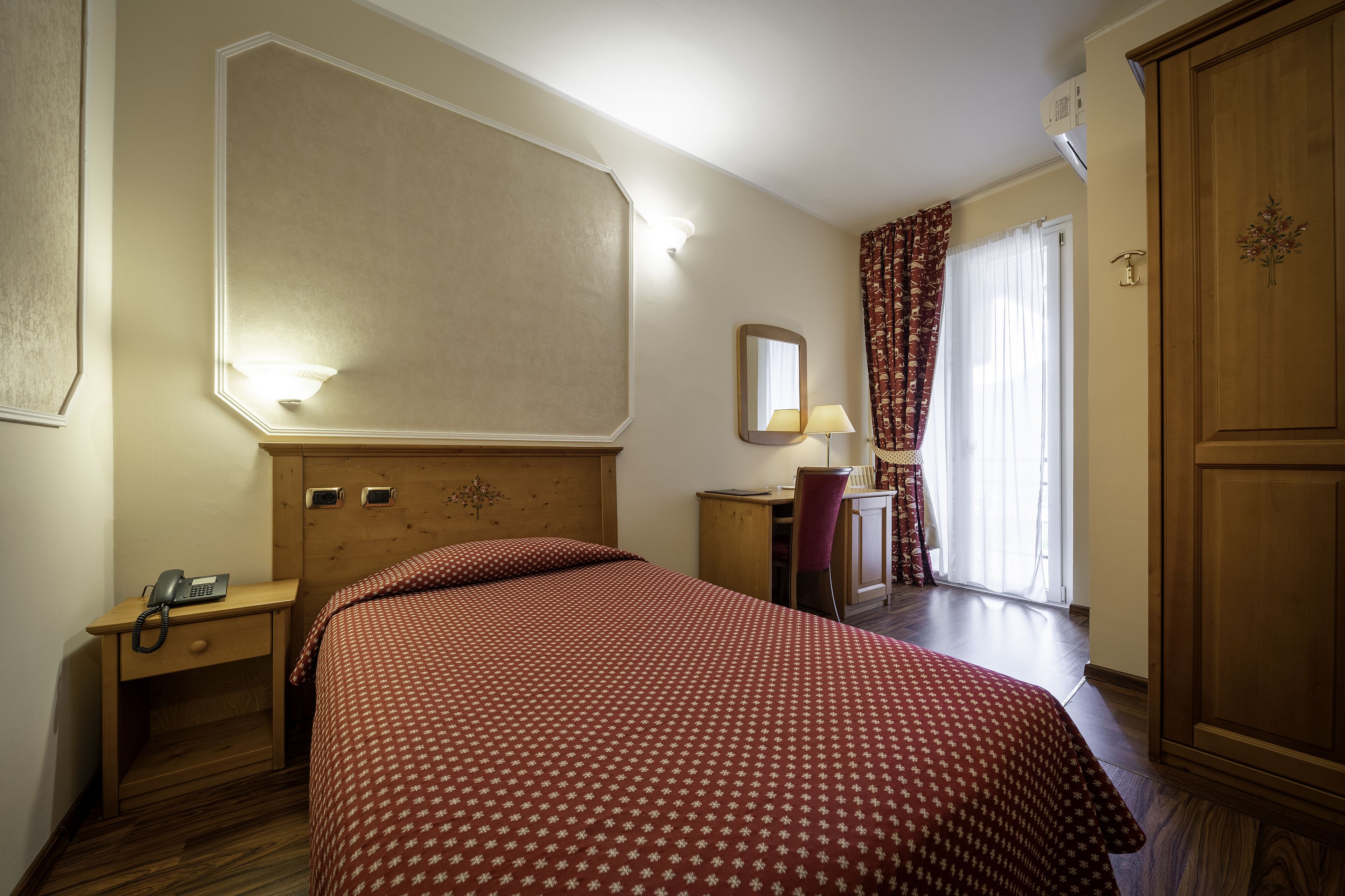 A hotel room with a bed, a desk with a lamp, a wardrobe, and a window with curtains.