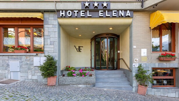 L’entrée de l’hôtel Elena se distingue par sa façade en pierre, une porte tournante en verre et des jardinières disposées sur les marches.
