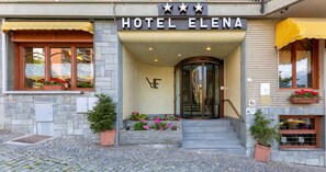 The entrance of Hotel Elena with a stone facade, a glass revolving door, and flower boxes on the steps. - Hotel Elena (Saint-Vincent)