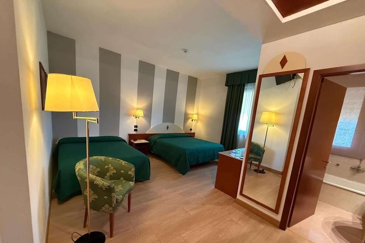 A hotel room with two beds, a chair, a desk, a mirror, and a bathroom with a shower.