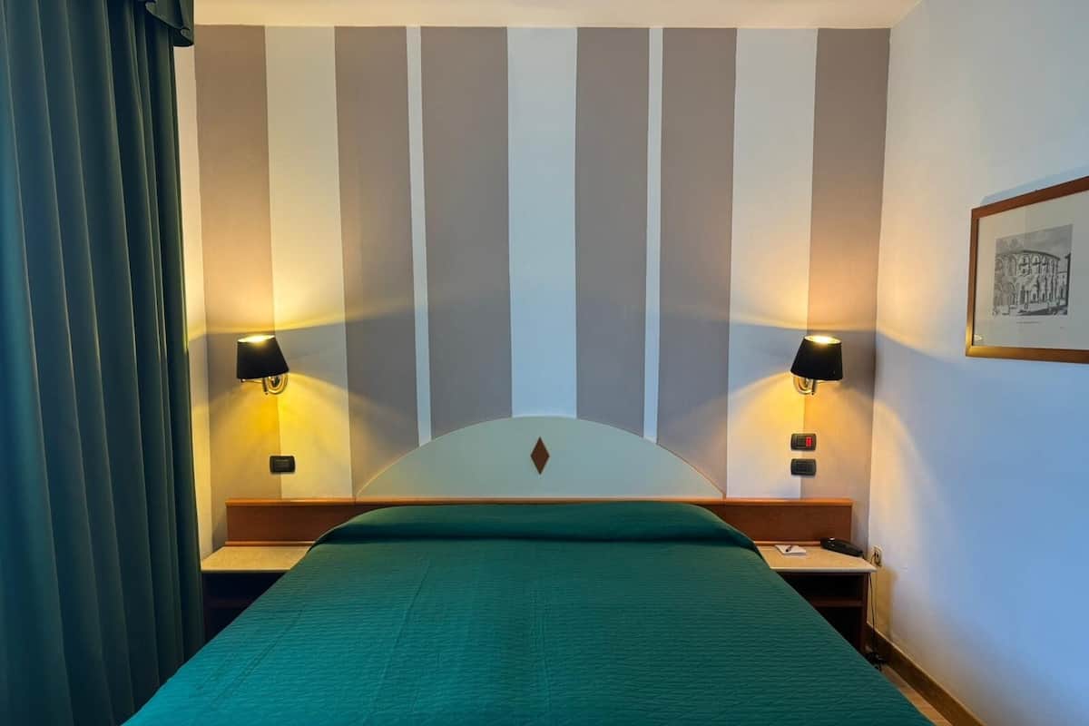 A hotel room with a bed, two bedside lamps, a headboard, and a framed picture on the wall.
