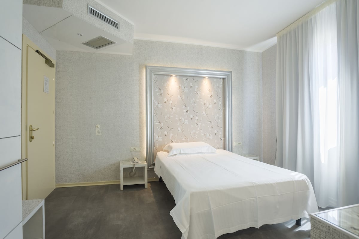 A hotel room with a bed, a nightstand, a small table, and a window with curtains.