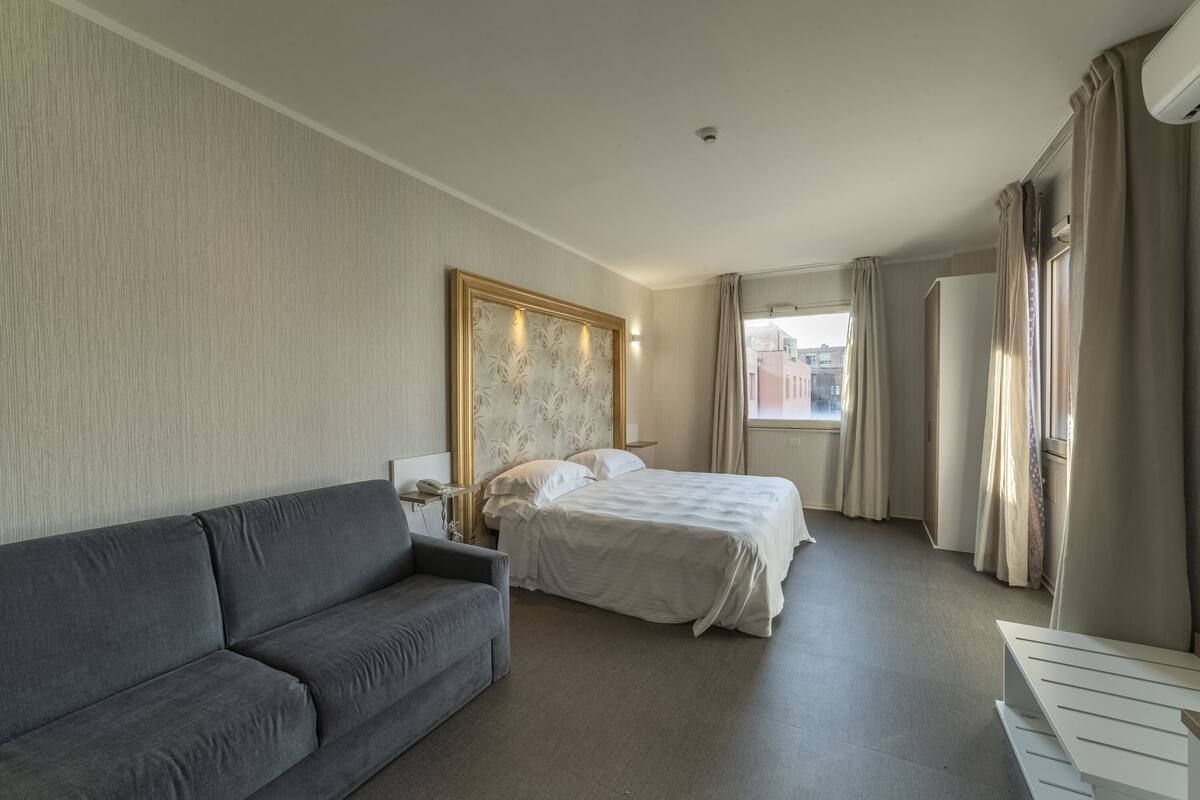 A hotel room with a gray sofa, a bed with white linens, a large framed artwork on the wall, and a view of buildings through the window.