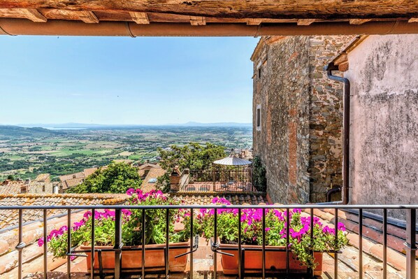Un balcon orné de pots en terre cuite remplis de fleurs, une vue sur un paysage rural et un bâtiment en pierre surmonté d’un toit de tuiles.