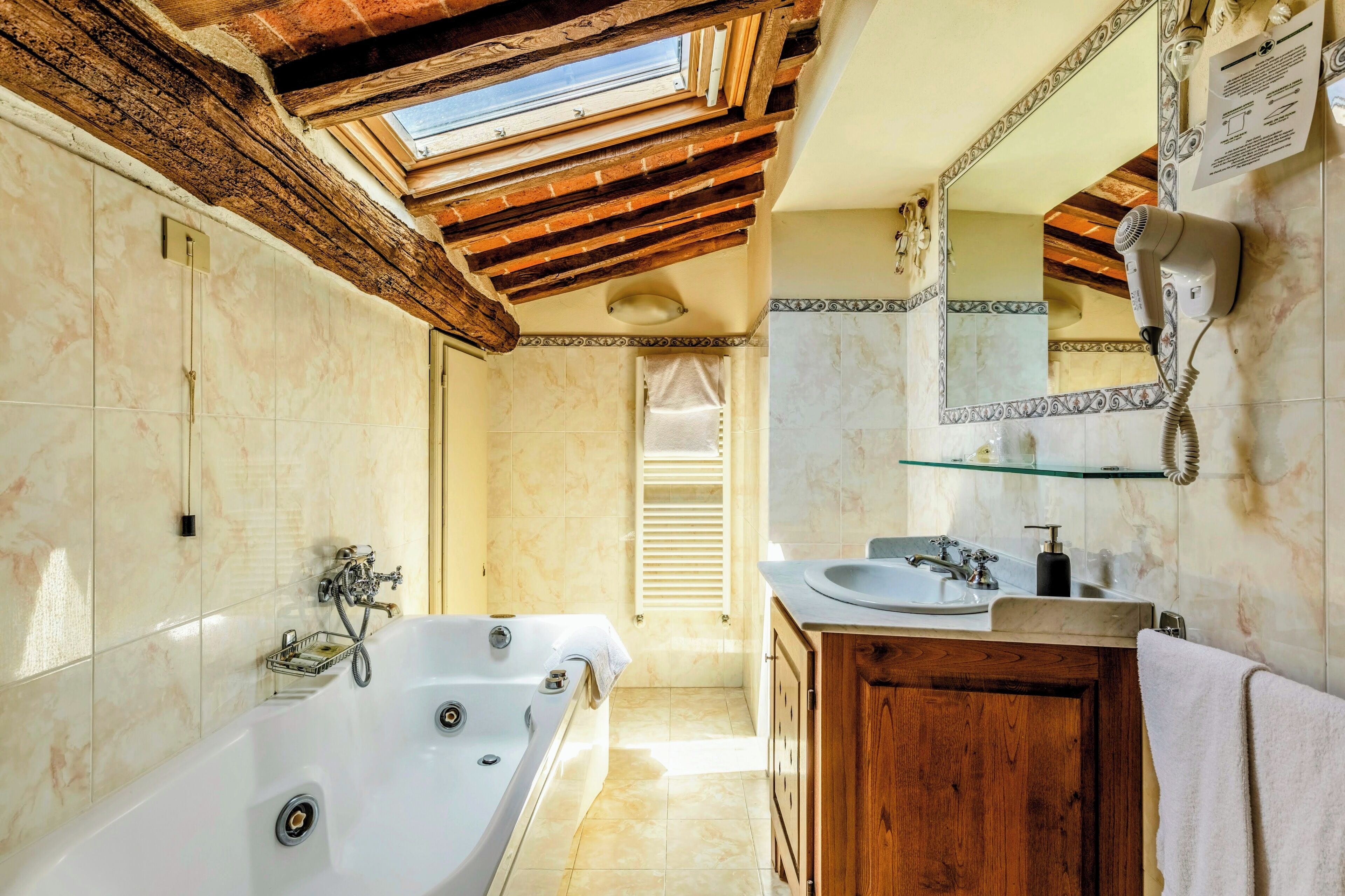 Un bagno con una grande vasca, travi in legno, un lucernario, uno scaffale in vetro e uno specchio con asciugacapelli.