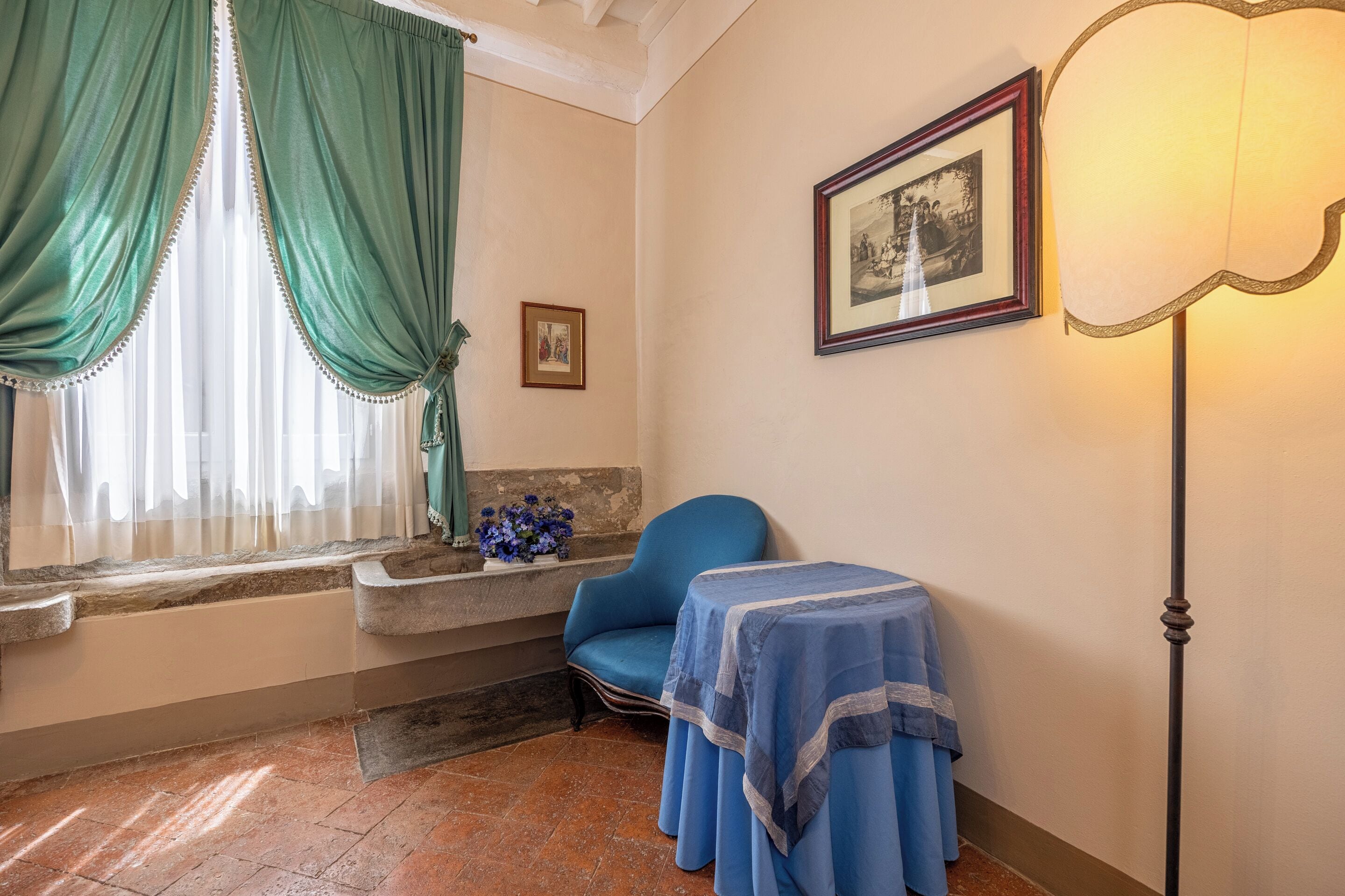 Ein Zimmer mit blauem Sessel, einem Tisch mit blauer Tischdecke, einem gerahmten Bild an der Wand und einem Fenster mit grünen Vorhängen.