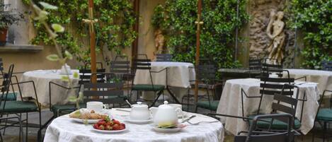 Un área de comedor al aire libre con mesas cubiertas con manteles blancos, sillas verdes y una estatua al fondo.