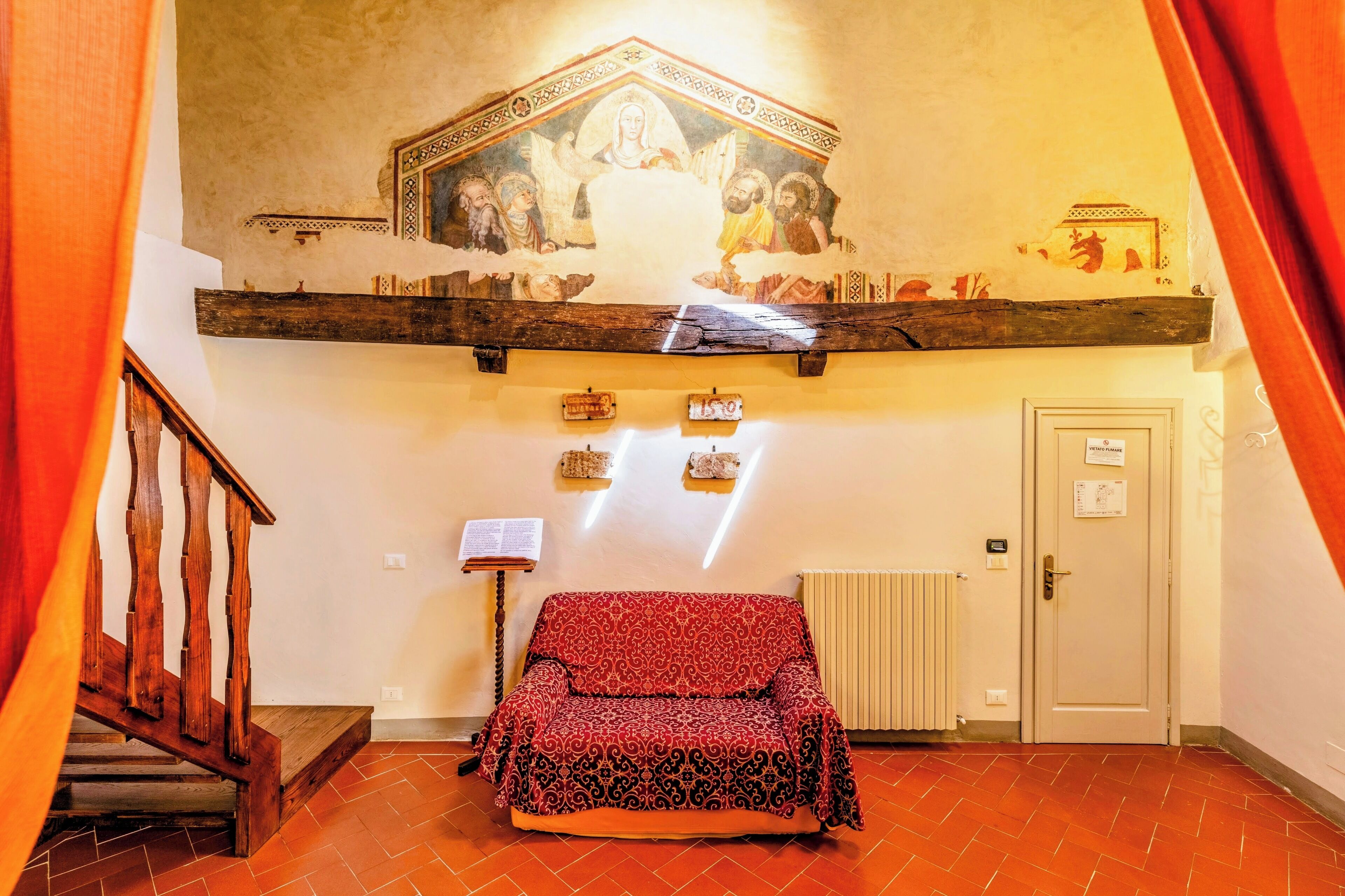 Une pièce avec un escalier en bois, un canapé rouge à motifs, une peinture murale au plafond et une porte avec une pancarte.