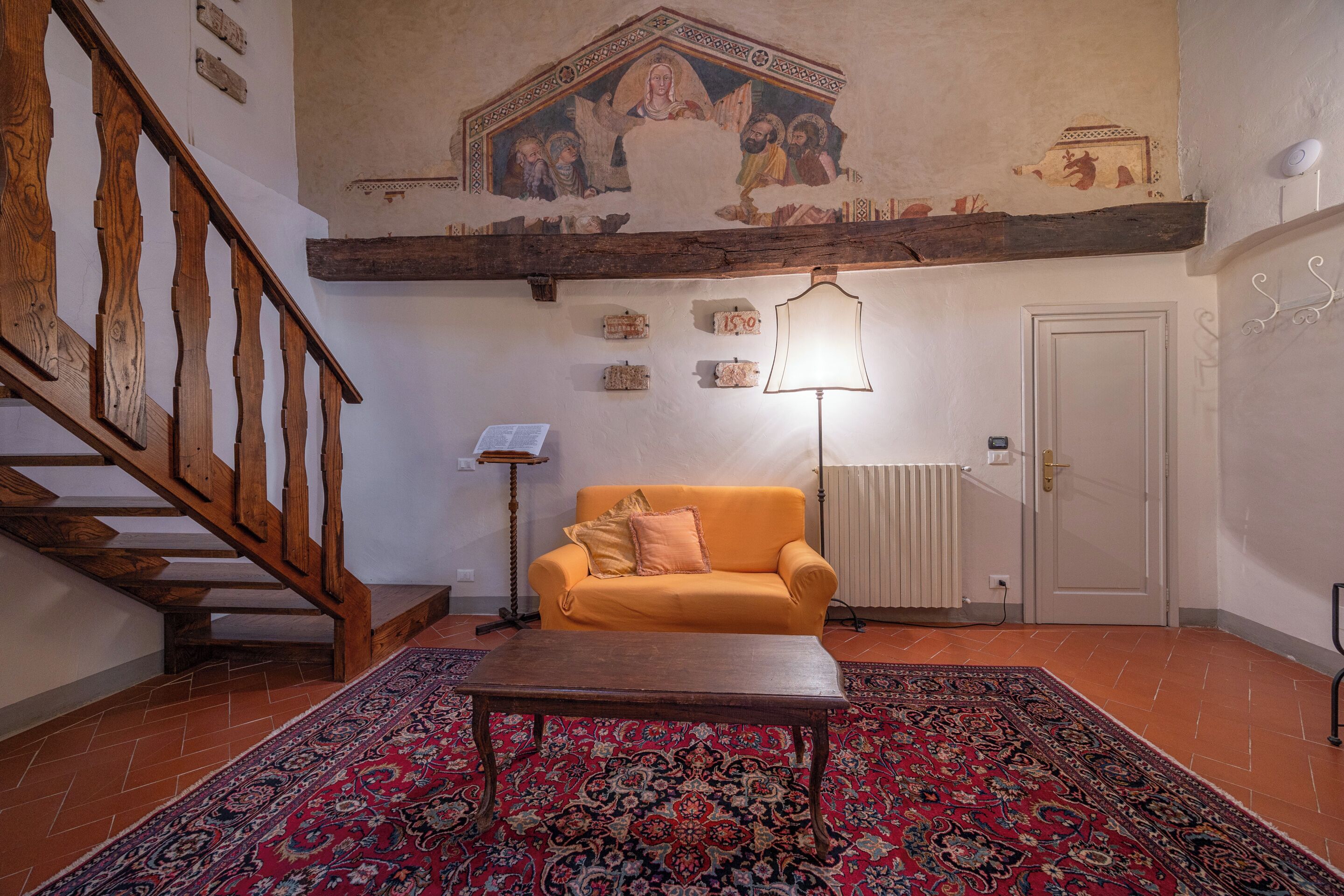 Una habitación con escalera de madera, un sofá, una mesa, alfombra estampada y un fresco en el techo.