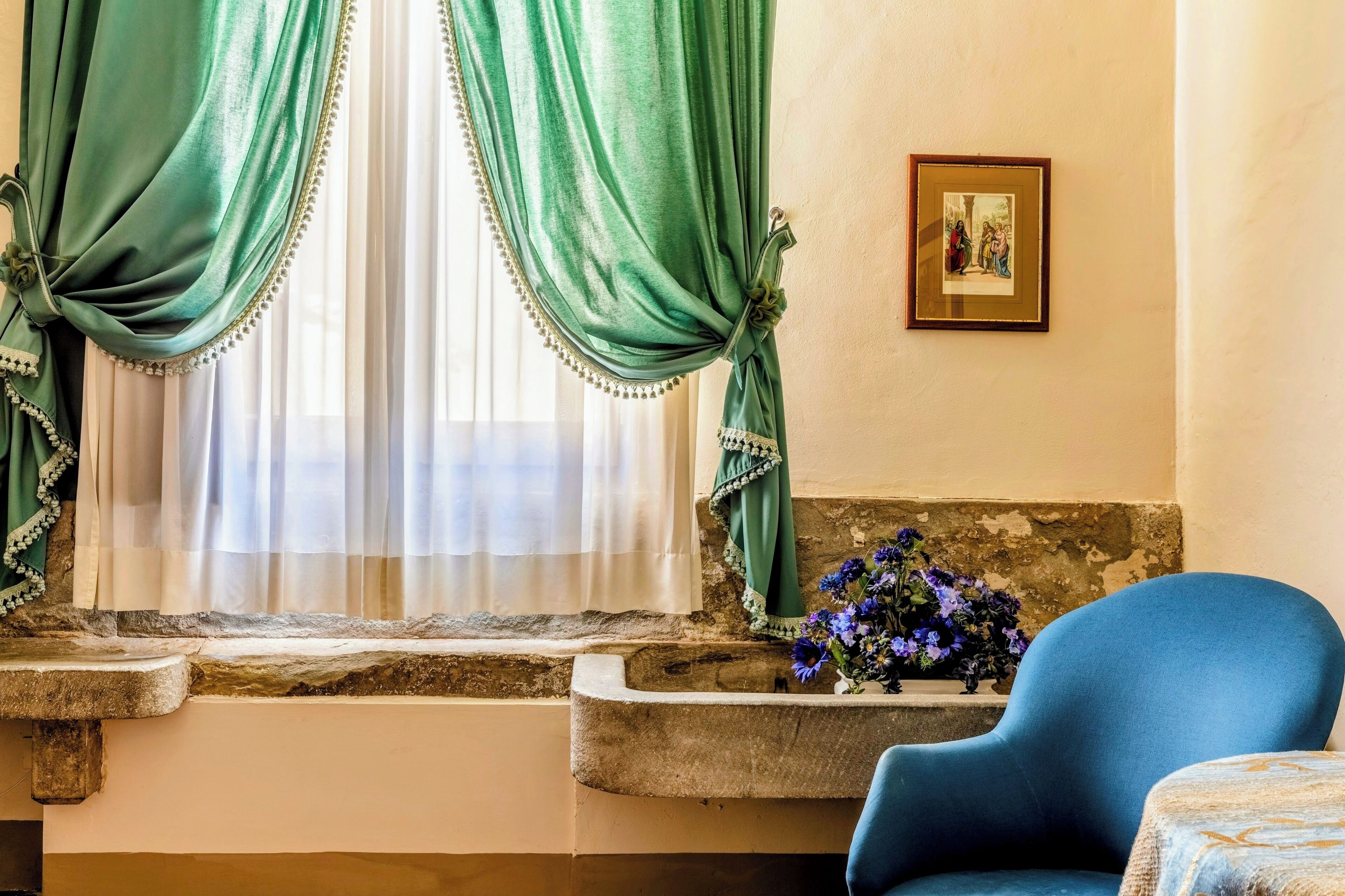 A room with a stone ledge, a blue chair, a vase with flowers, and a framed picture on the wall.