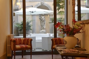 Quarto com duas poltronas estofadas, uma mesa com revistas, um vaso grande com flores e vista para área externa com estátua e plantas.