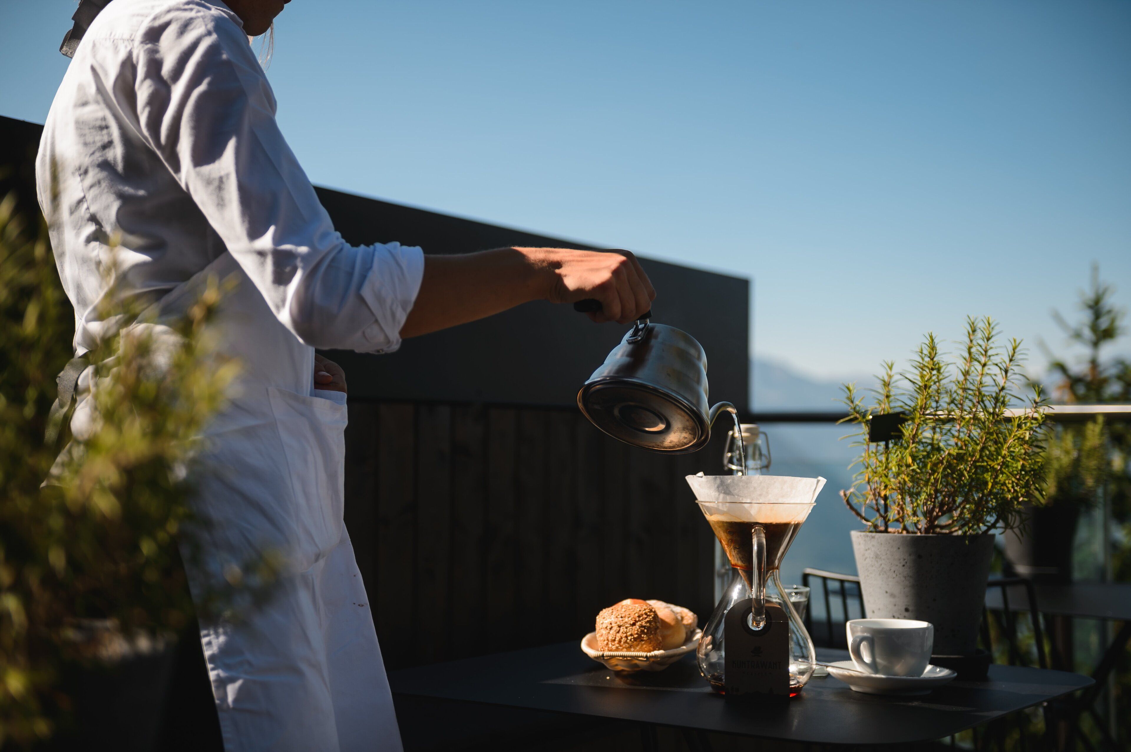 A person pouring coffee into a cup on a balcony with a view of the sea and a potted plant.