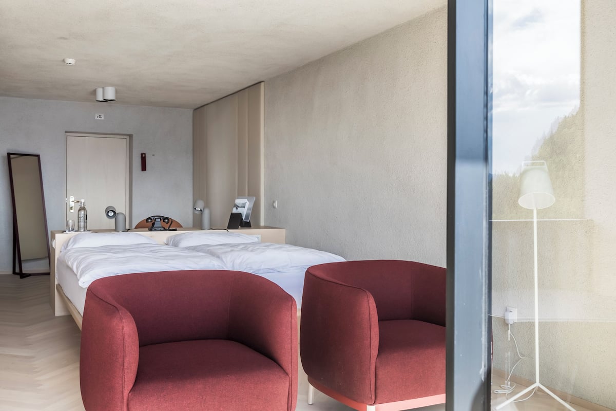 Ein modernes Hotelzimmer mit einem Bett, zwei roten Sesseln, einem Nachttisch mit Lampe und einem großen Spiegel an der Wand.