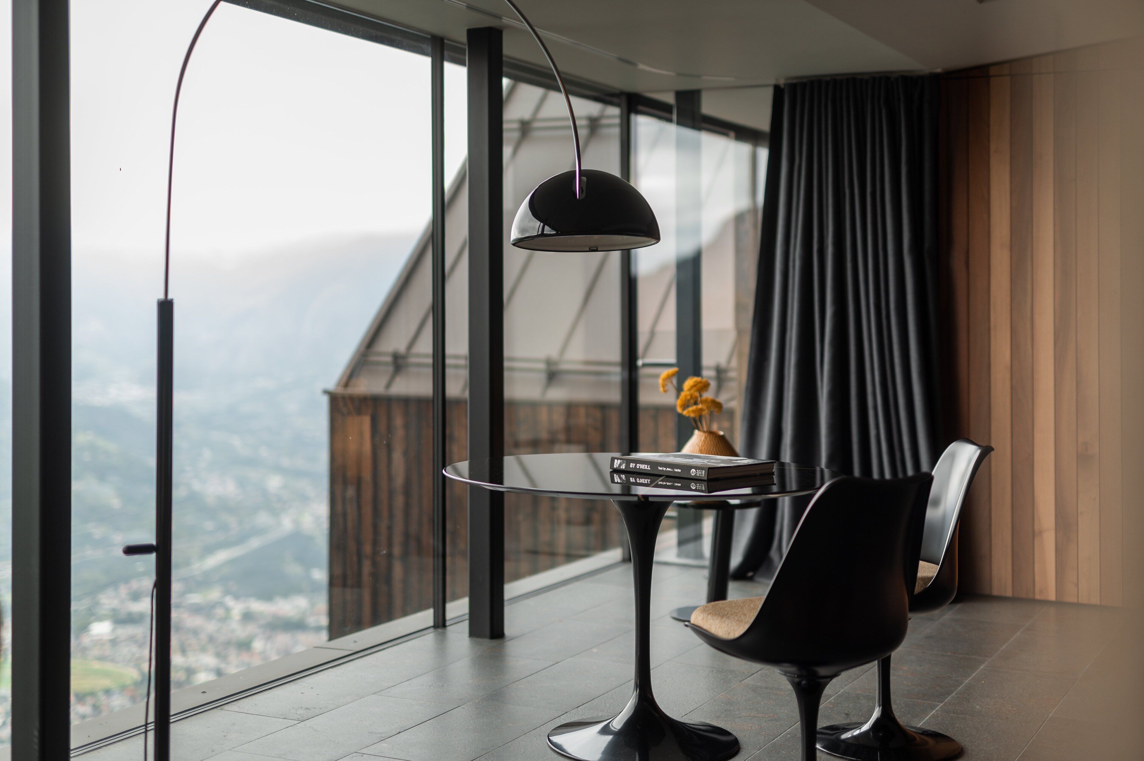 A modern room with a large window, a round table with a book, a chair, and a pendant light.