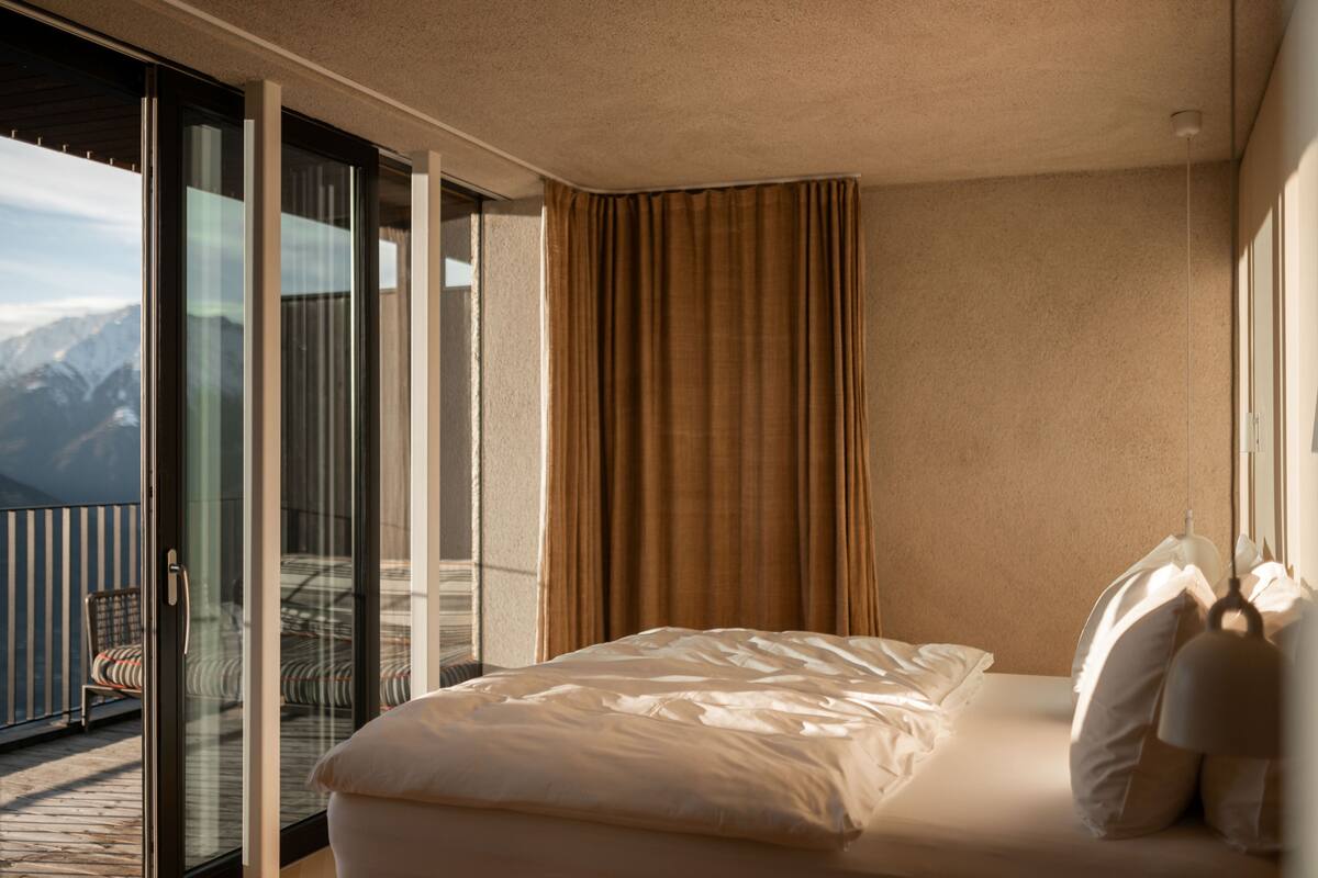 Ein Hotelzimmer mit einem großen Bett, Blick auf Berge durch eine Schiebetür und einem Balkon mit einem Sessel.