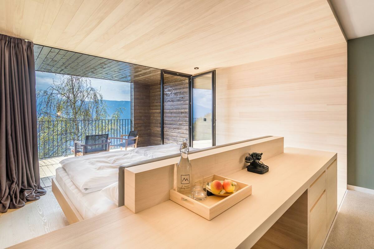 Ein modernes Schlafzimmer mit einem großen Bett, einem Holztisch mit Obstkorb und Blick ins Freie.