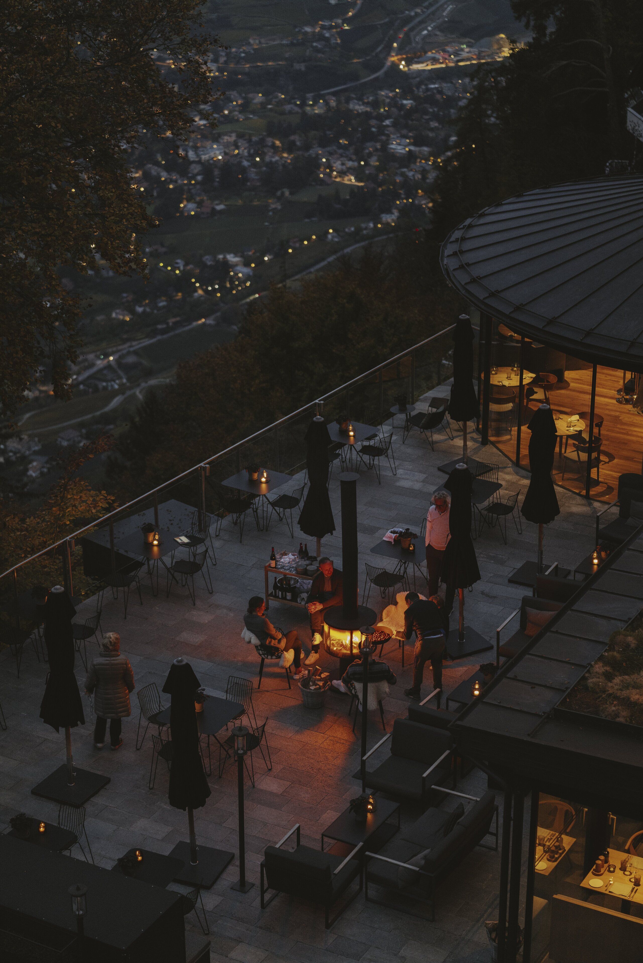 An outdoor patio area with tables and chairs, a fire pit, and a view of a town below.
