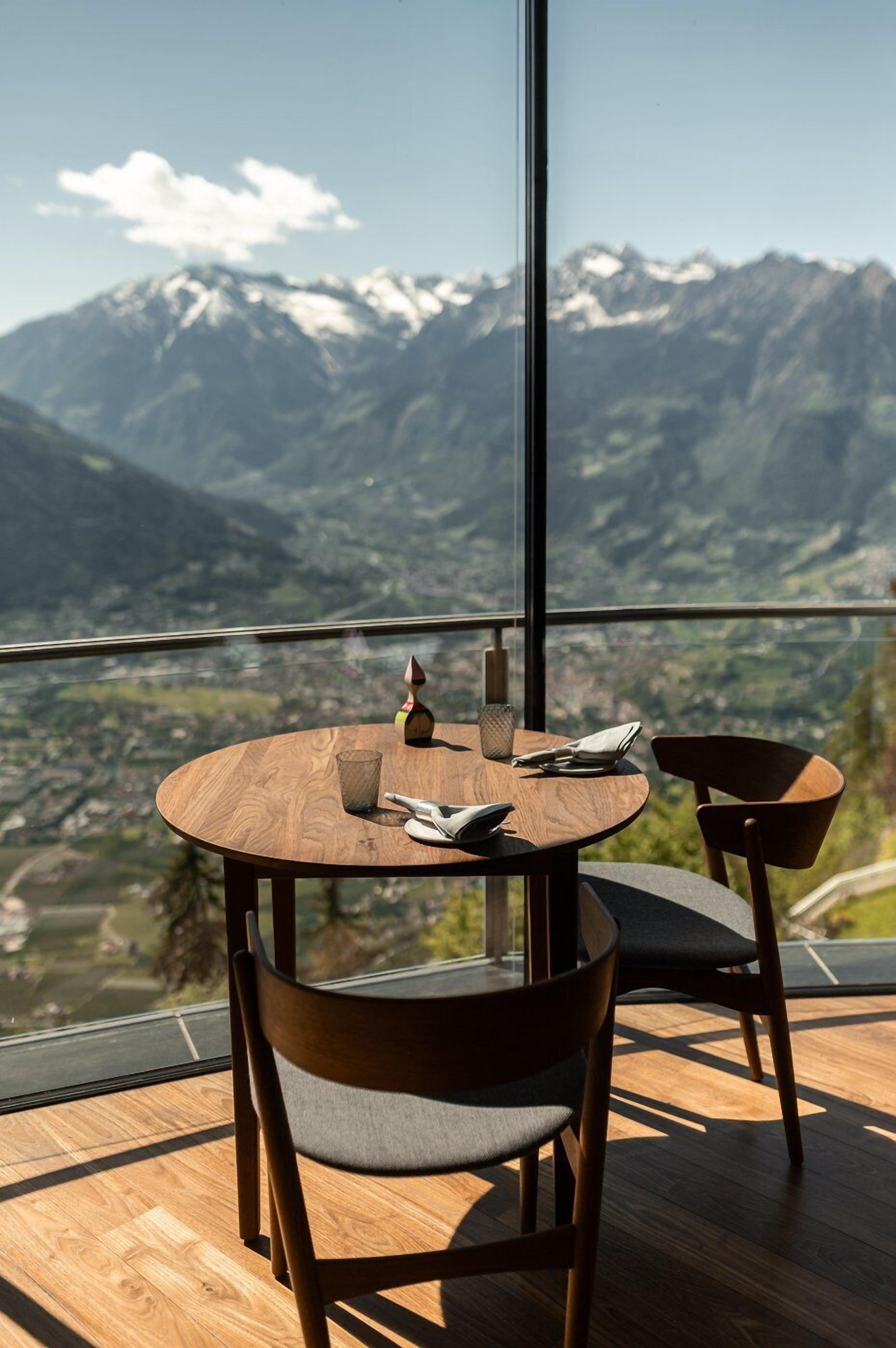 A round wooden table with two chairs, a glass, and a napkin on a balcony with a mountain view.