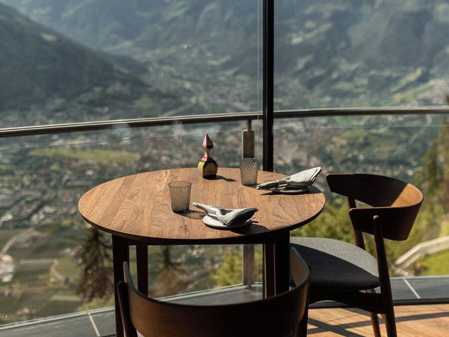 A round wooden table with two chairs, a glass, and a napkin on a balcony with a mountain view.