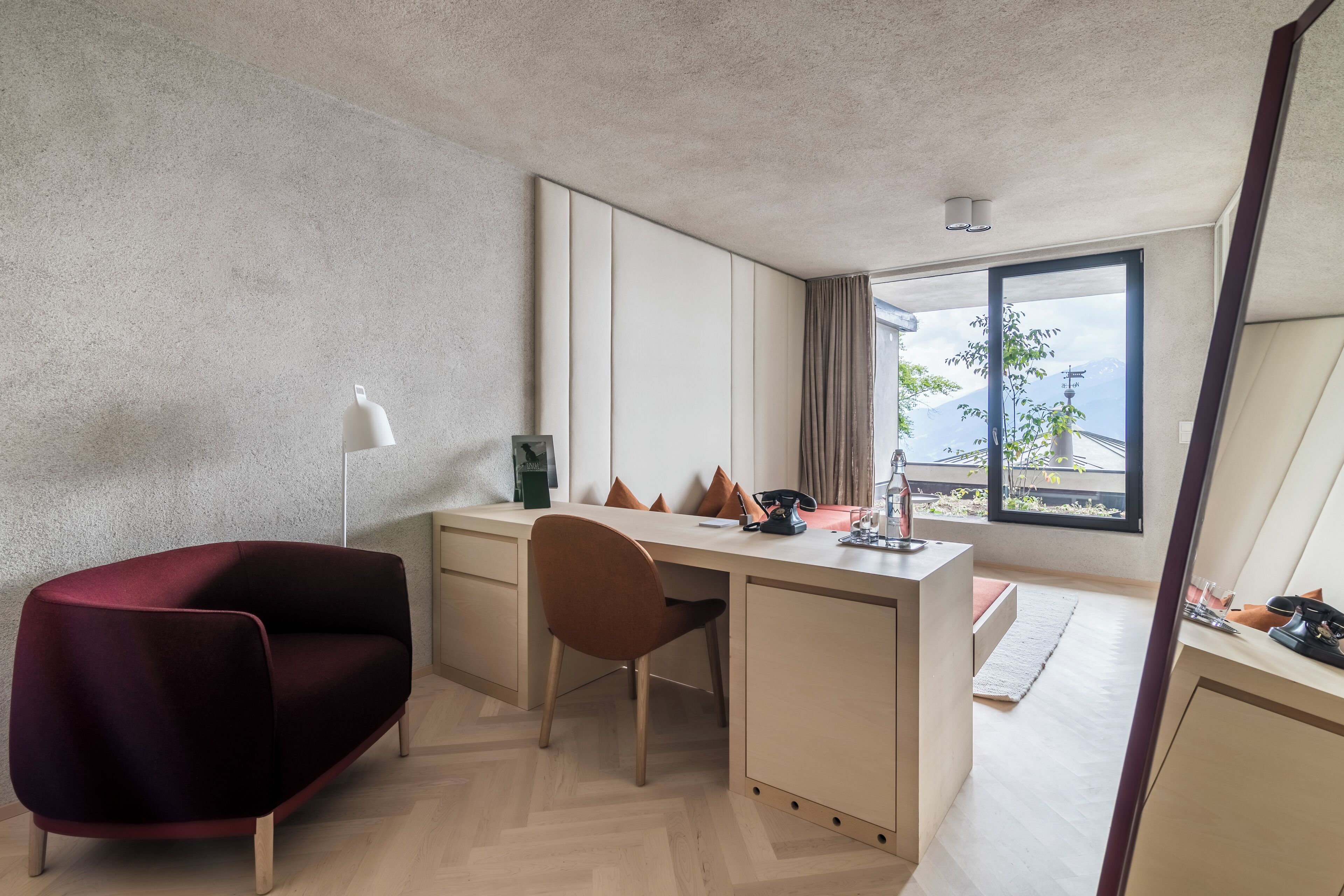 A modern hotel room with a desk, a red sofa, a chair, and a large window offering a view of the outdoors.
