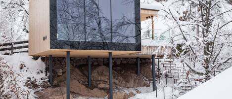 A modern cabin with a large glass window, wooden exterior, and a stone foundation.