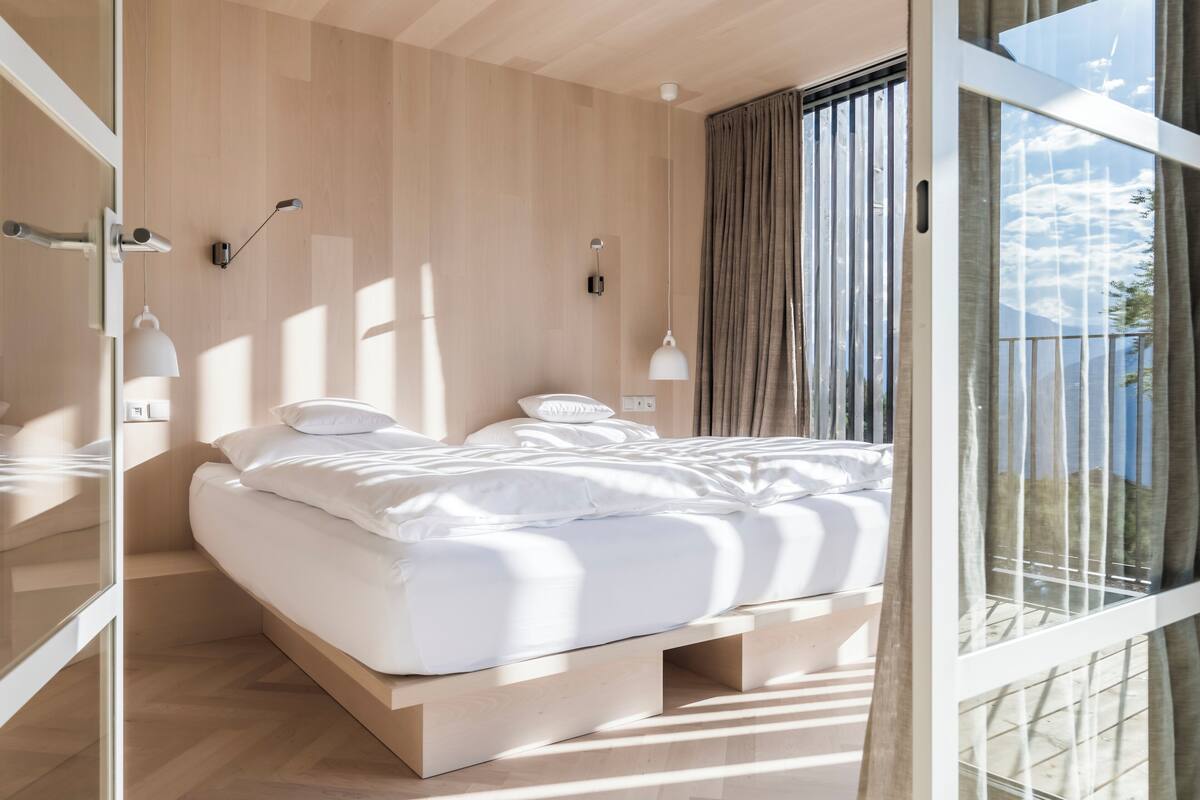 Ein modernes Schlafzimmer mit Holzboden, einem Bett mit weißer Bettwäsche und einem großen Fenster mit Vorhängen.