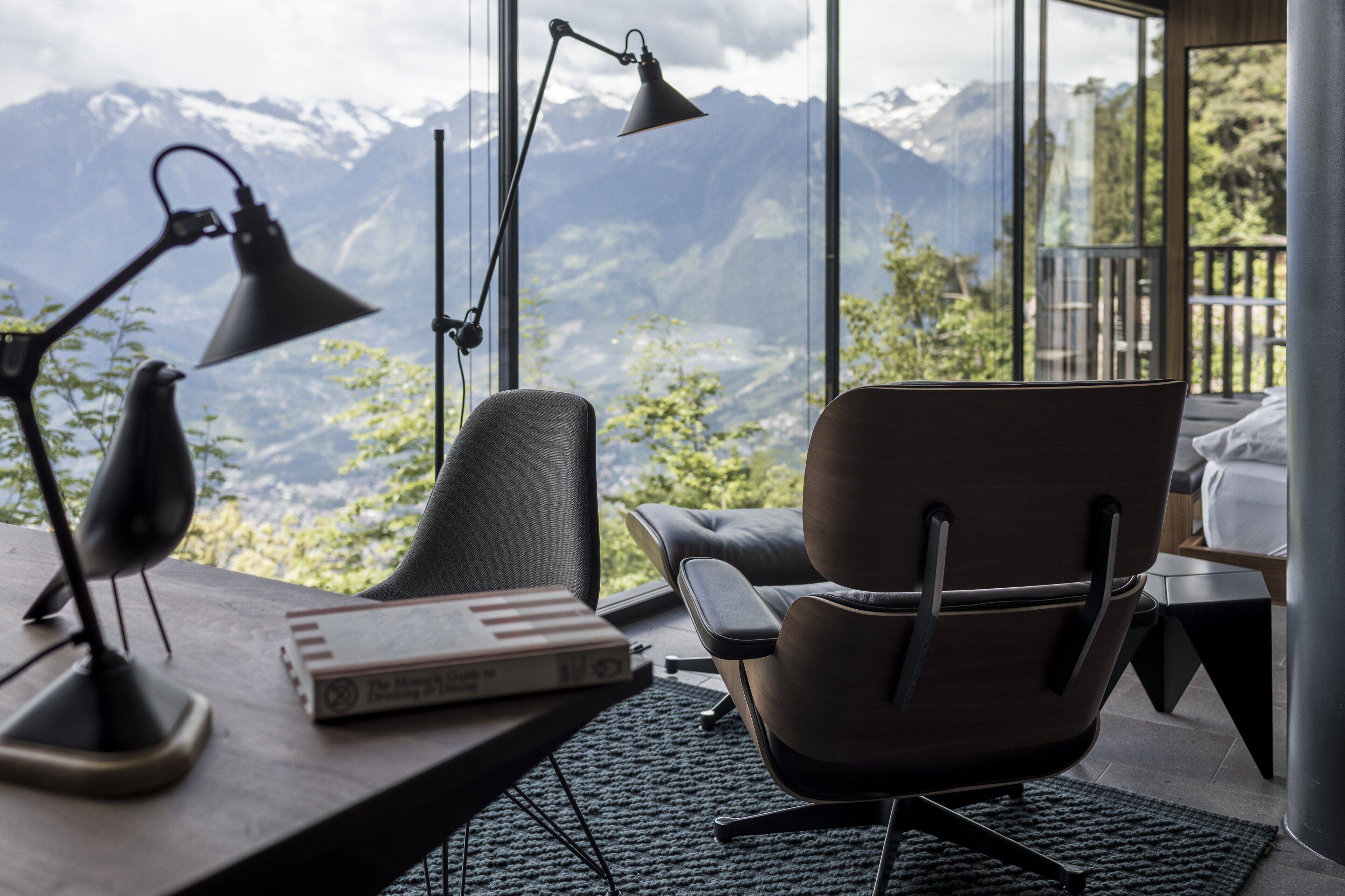 Una habitación moderna con vistas a las montañas, un escritorio con lámpara, una silla y un libro.