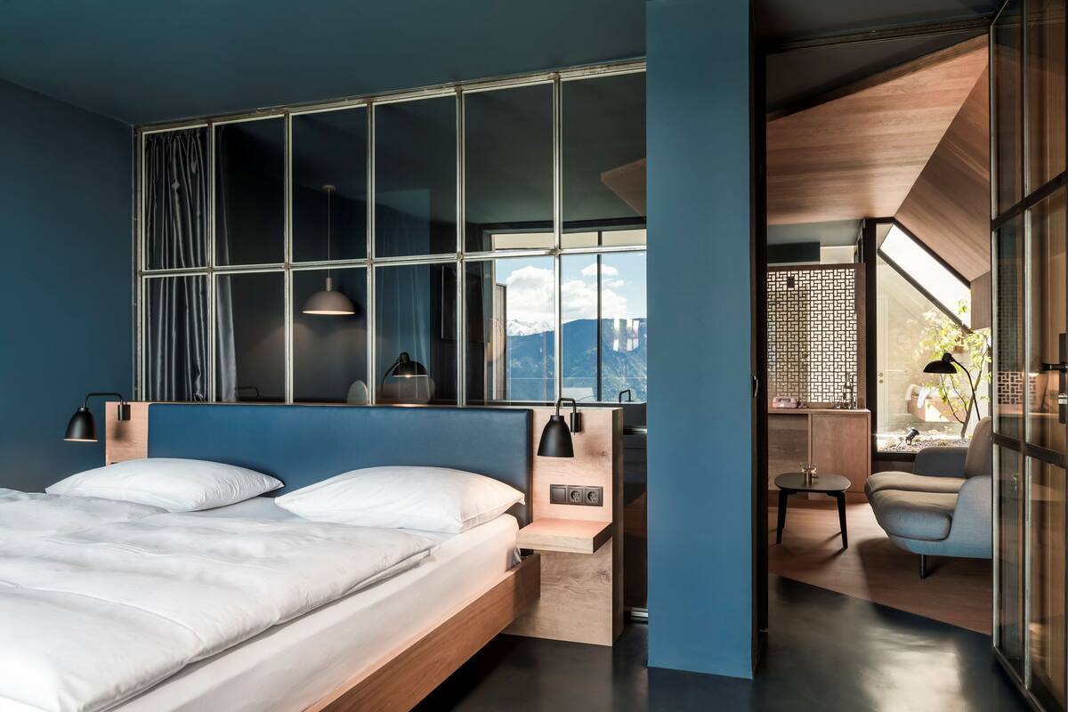 Ein modernes Schlafzimmer mit einem großen Bett, Nachttischlampen, einem kleinen Tisch und Bergblick durch große Fenster.