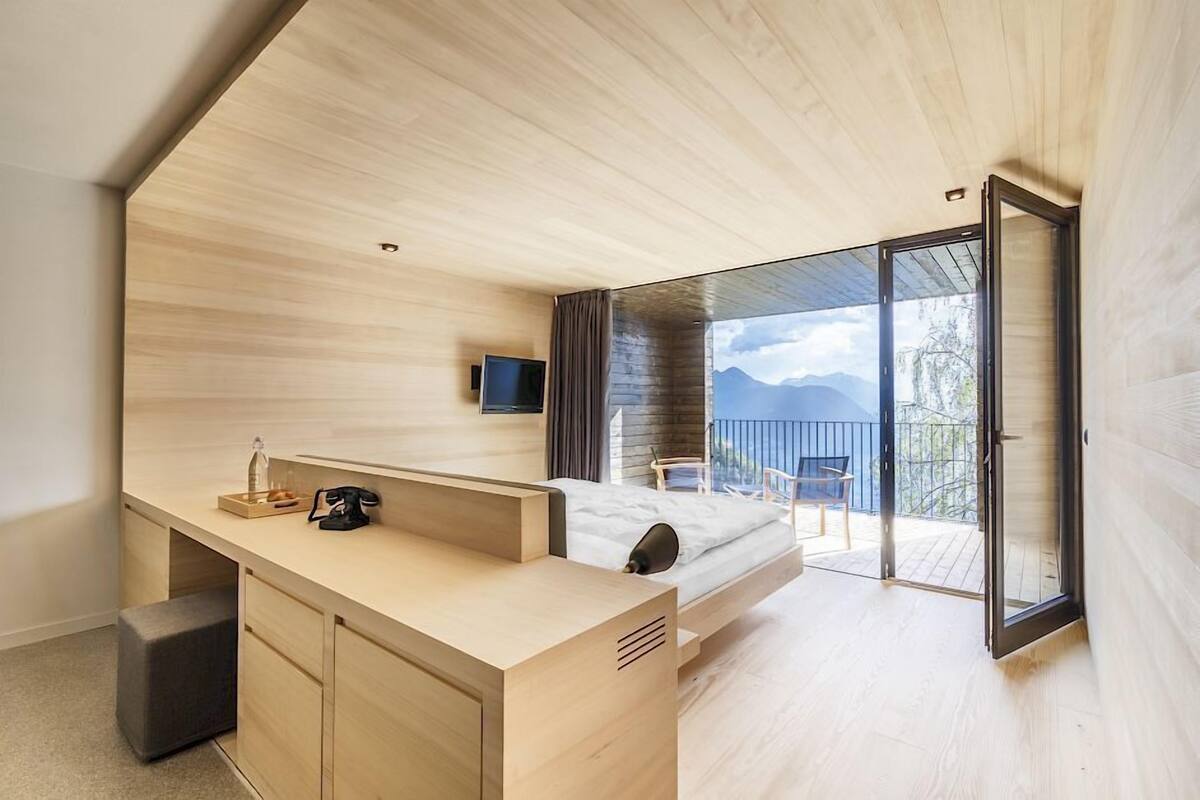 Ein modernes Schlafzimmer mit Holzboden, einem Bett, einem Schreibtisch, einem Fernseher und einem Balkon mit Bergblick.