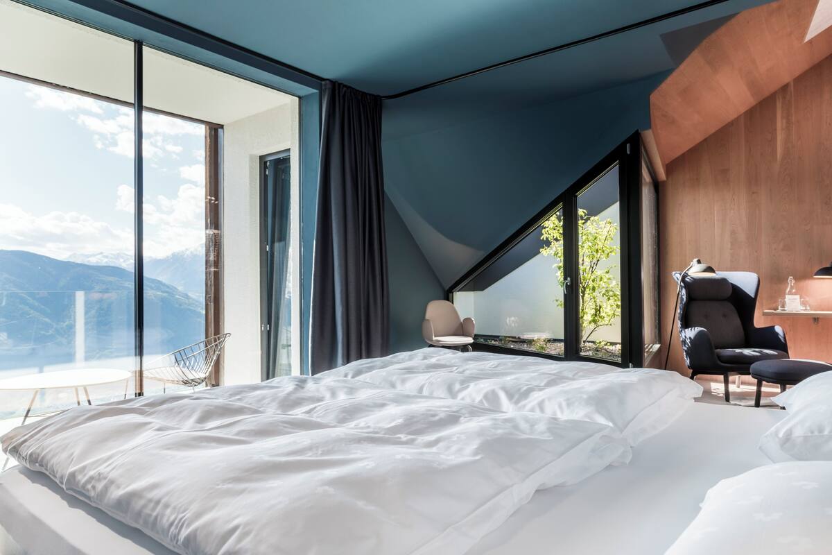 Ein modernes Hotelzimmer mit einem großen Bett, einem Schreibtisch und einem Stuhl. Durch große Fenster ist eine Bergblick zu sehen.