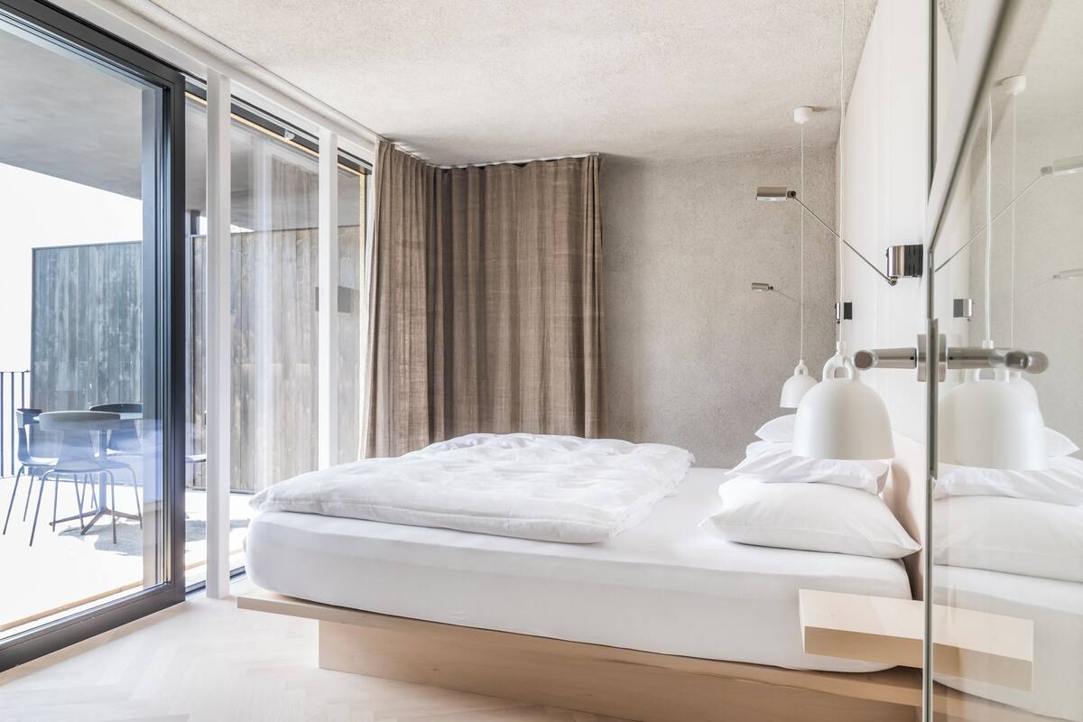 Ein modernes Hotelzimmer mit einem großen Bett, einer Glastür zu einem Außenbereich und einem minimalistischen Design.