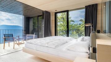 A modern bedroom with a large bed, a balcony with a view of the mountains, and a seating area with chairs.