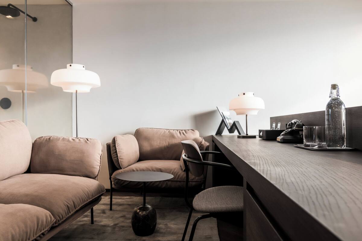 Ein modernes, minimalistisches Wohnzimmer mit einer Couch, einem kleinen runden Tisch und zwei Pendelleuchten. Ein Holztisch mit einem Laptop und einer Wasserflasche ist sichtbar.
