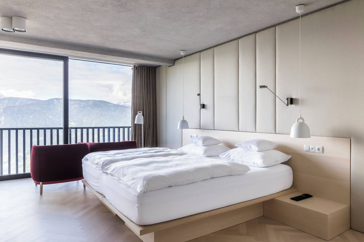 Ein modernes Hotelzimmer mit einem großen Bett, einem roten Sessel, einem Nachttisch und Bergblick durch große Fenster.