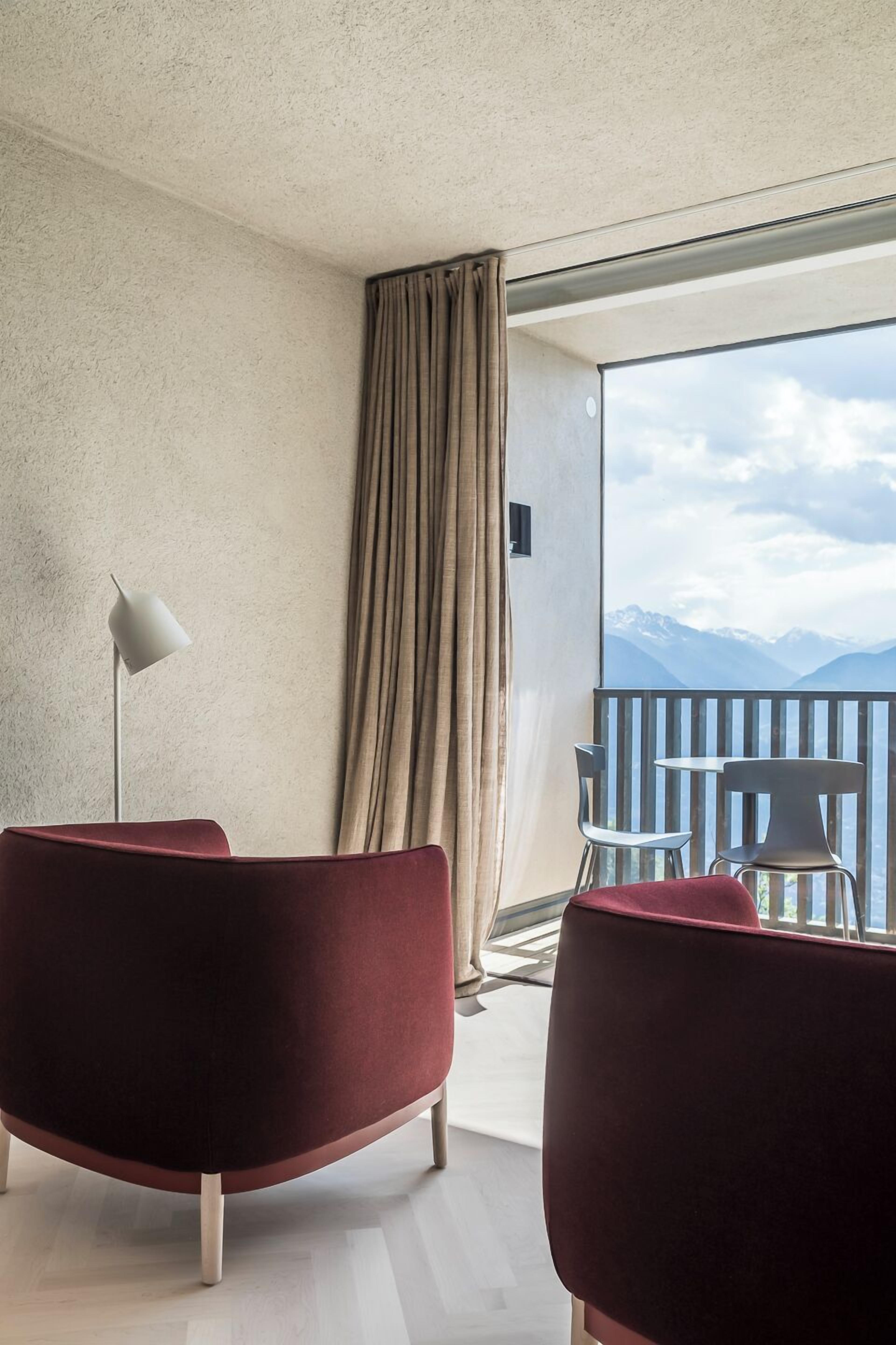 A hotel room with a large window, a red chair, a white floor lamp, and a view of mountains.