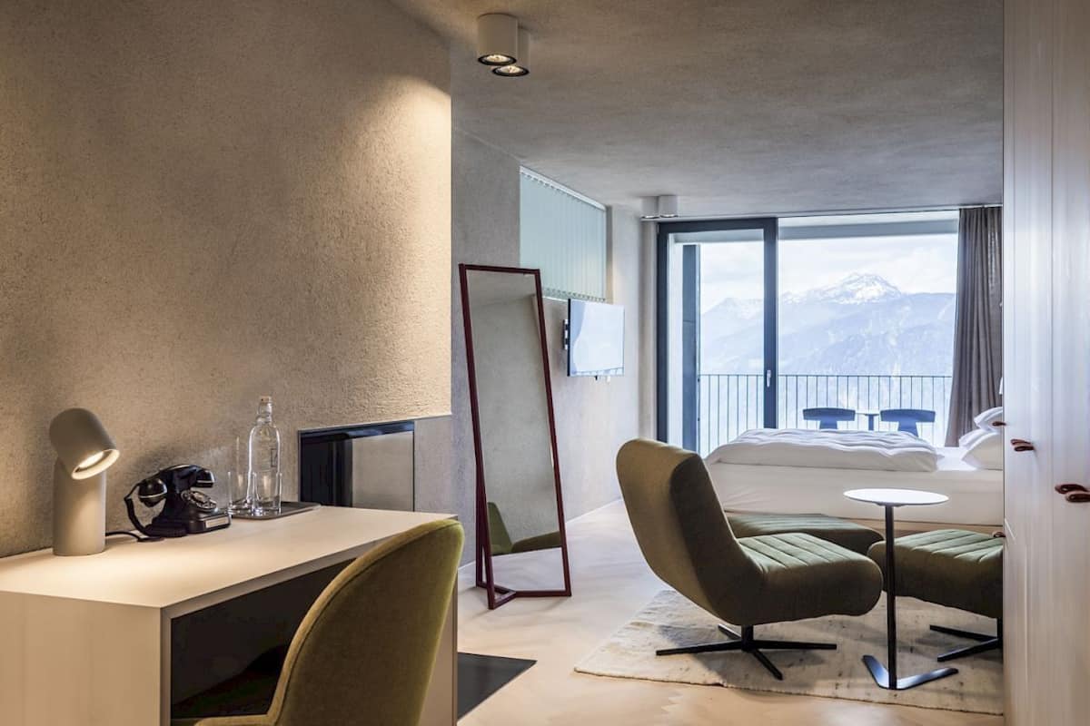 Ein Hotelzimmer mit Schreibtisch, Stuhl, Bett, großem Fenster mit Bergblick und einem Balkon mit Tischen und Stühlen.