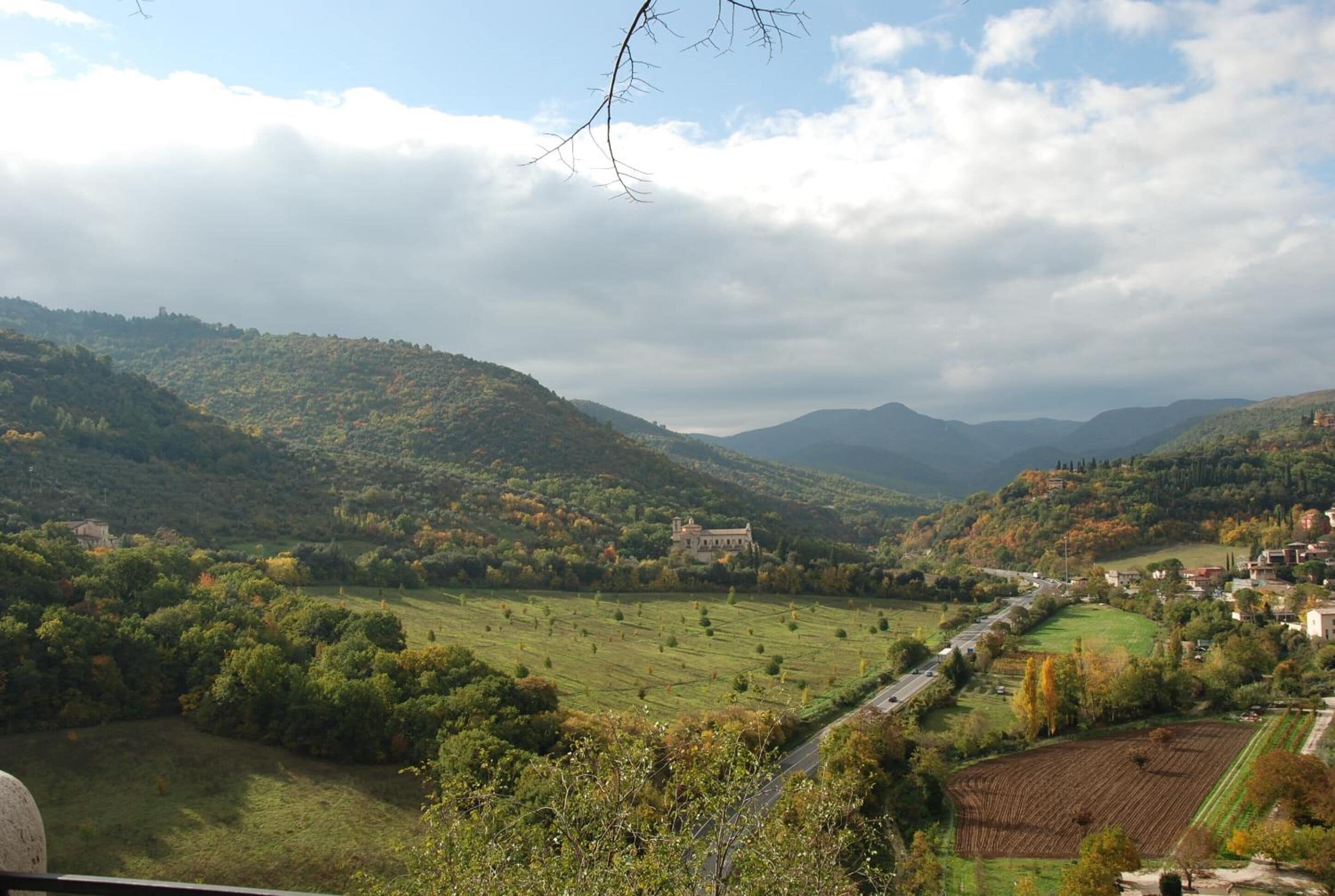 A panoramic view of a valley with a prominent building on a hill, surrounded by lush greenery and fields.