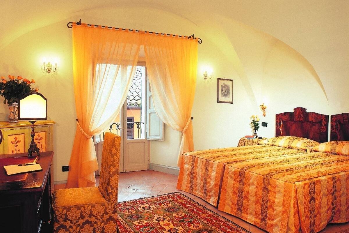 A bedroom with a large bed, a desk with a lamp, and a chair. There is a window with curtains and a view of the outside.