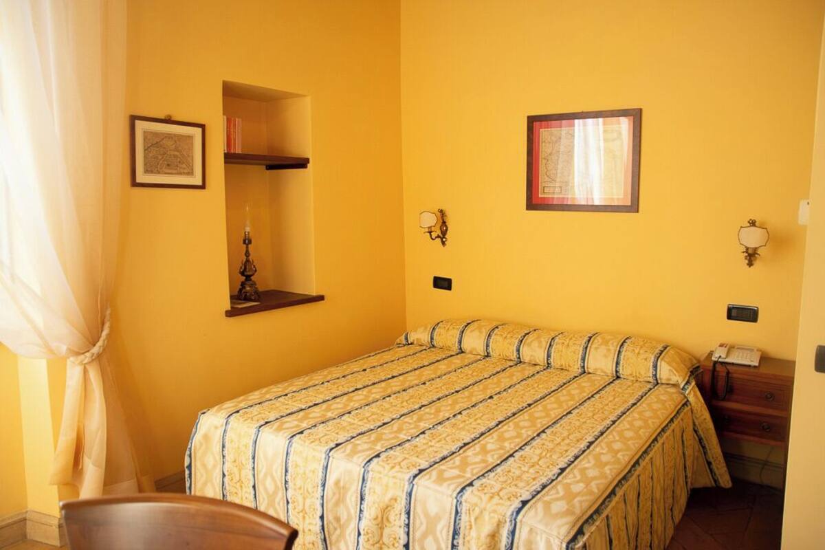 A room with a bed, a chair, a bedside table with a lamp, a shelf with books, and a framed picture on the wall.