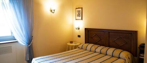 A bedroom with a bed, a bedside table, a nightstand, a window with curtains, and wall-mounted lights.