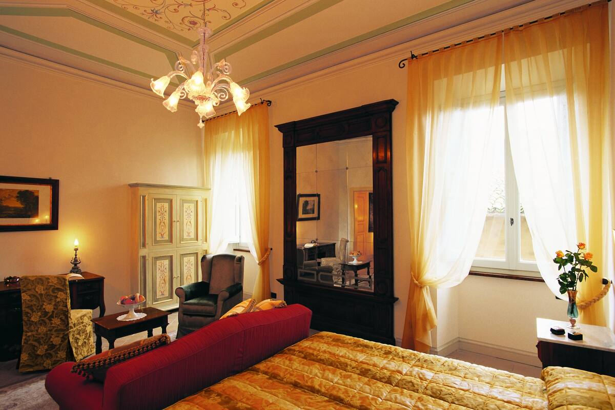 A hotel room with a large bed, a sitting area with a red sofa, a wooden cabinet, a chair, and a large mirror.