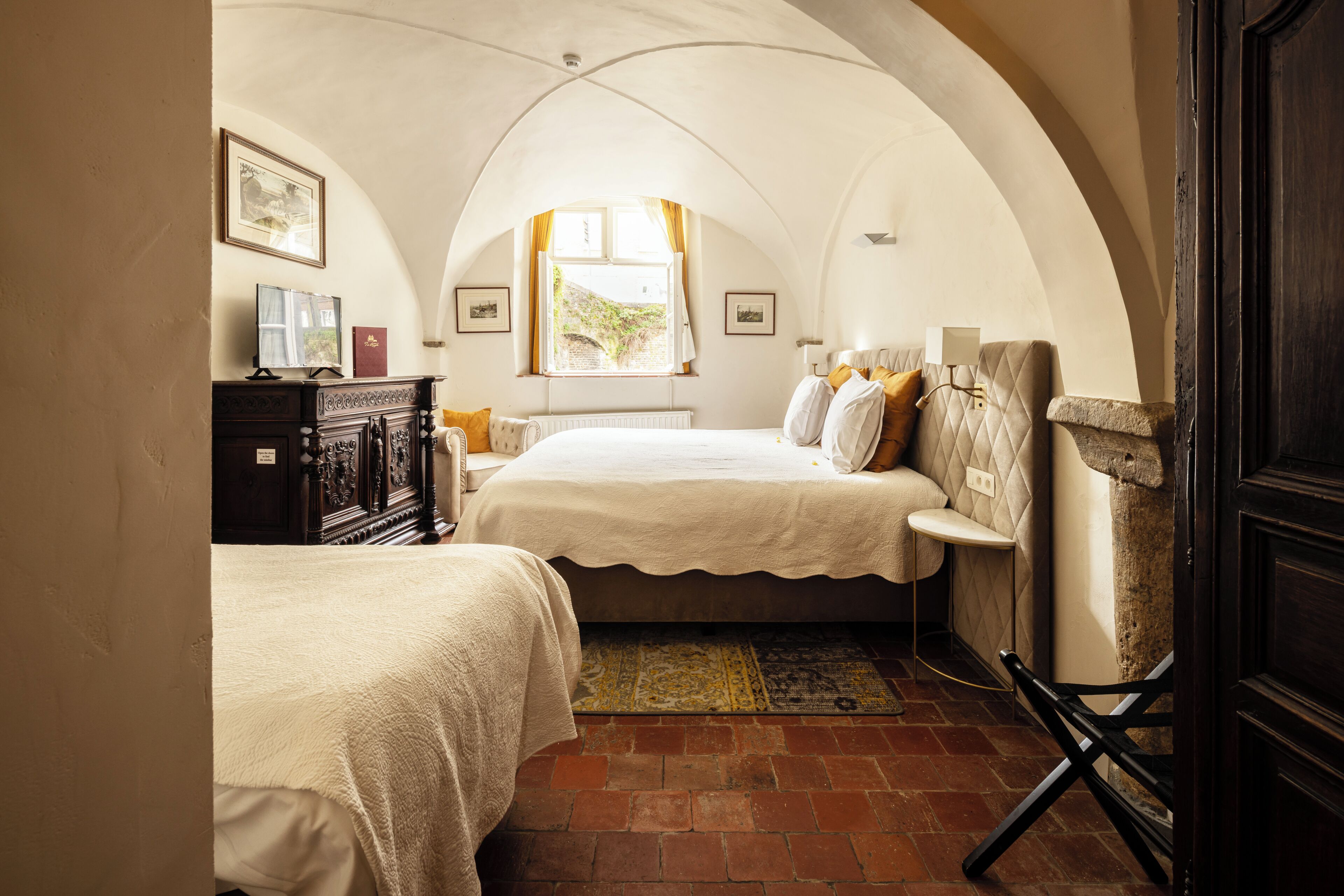 A bedroom with two beds, a dresser, a window with curtains, and a stone bench.