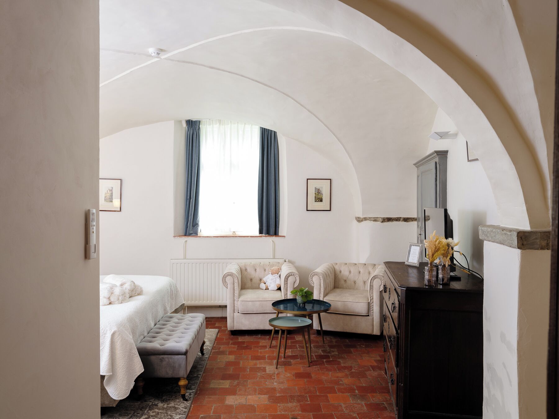 A cozy room with a bed, a sofa, a small table, and a dresser. There is a window with curtains, a radiator, and a mirror on the wall.